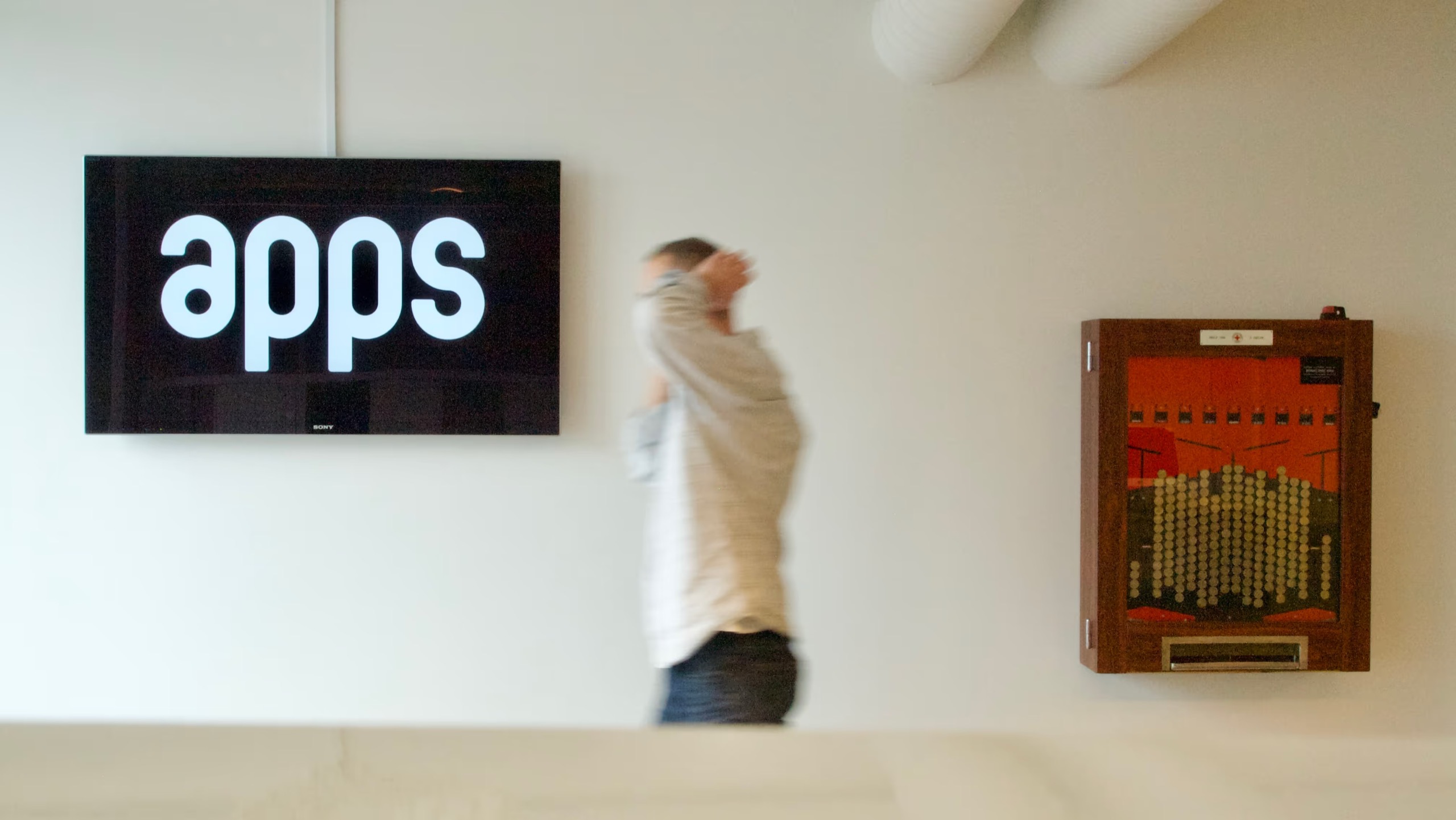 The Apps logo on a screen in the Oslo office, a colleague walking past, and the wall-mounted Kronespillet coin machine