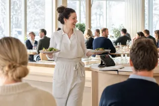 Servitør bærer to tallerkener gjennom et fullsatt restaurantlokale mens en kontortelefon på disken lyser rødt av et ubesvart anrop