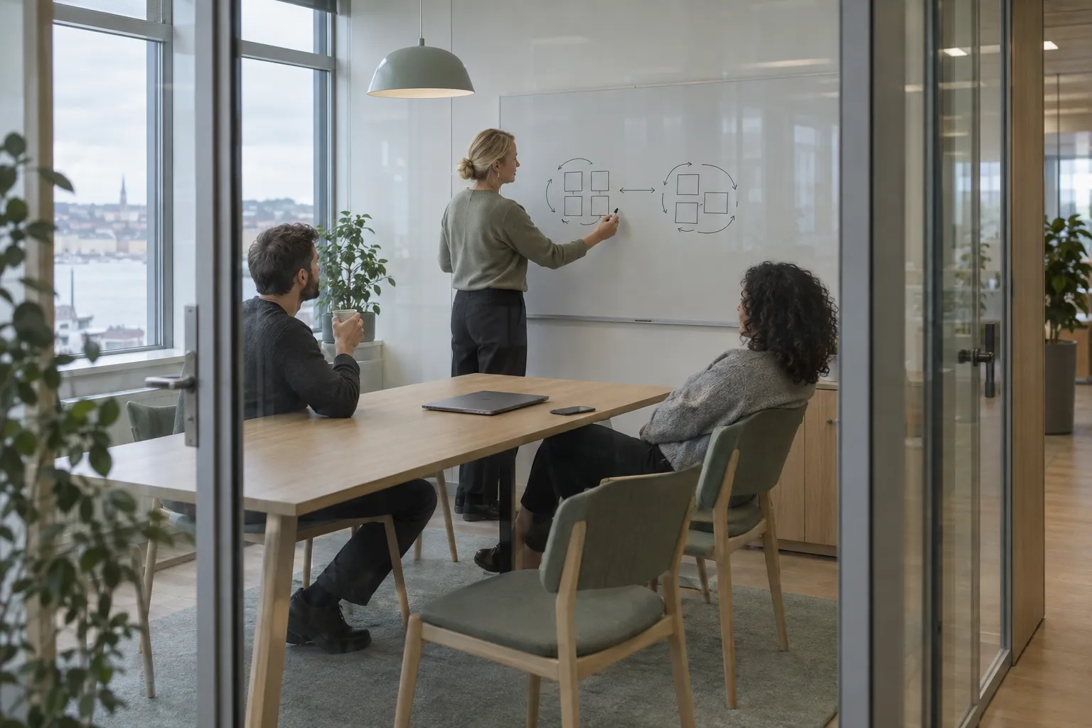 A glass-walled meeting room where a woman sketches two groups of boxes connected by arrows on a whiteboard while two colleagues listen at an oak table.