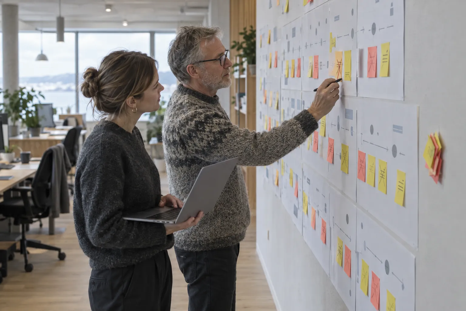 An older man in a knitted sweater marks a sticky note on a wall-sized process map of printed sheets and colored notes, while a younger woman beside him holds an open laptop.