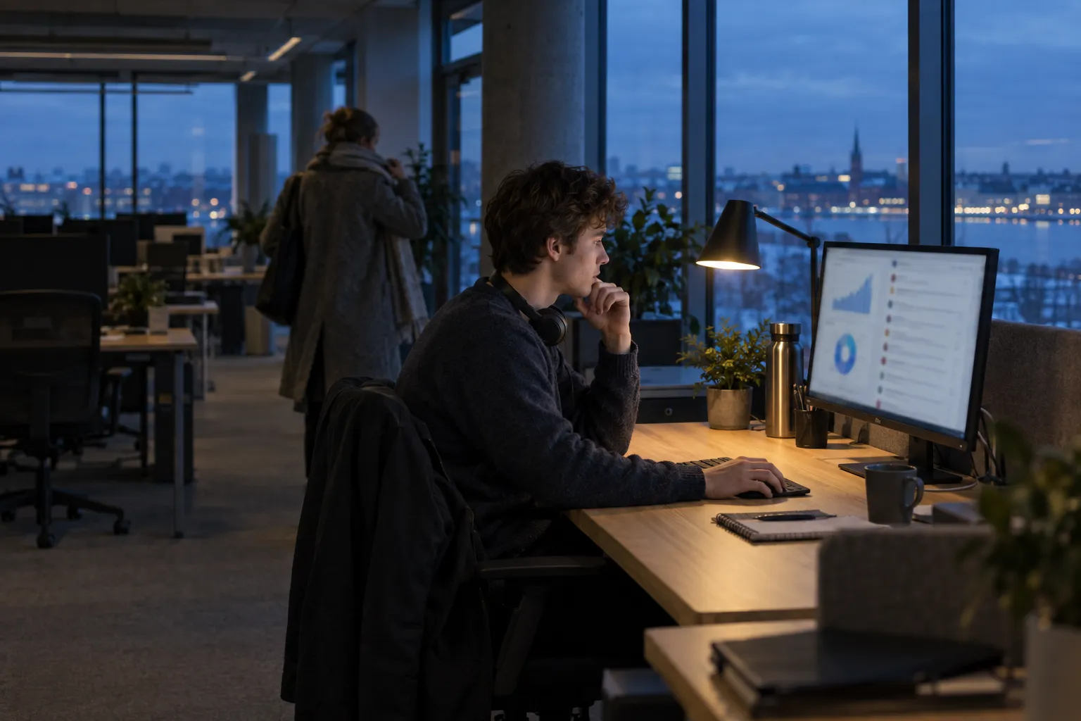 A dim open-plan office at dusk by tall windows with city lights outside, where a young man works under a desk lamp at a monitor showing charts while a colleague in a coat heads home.