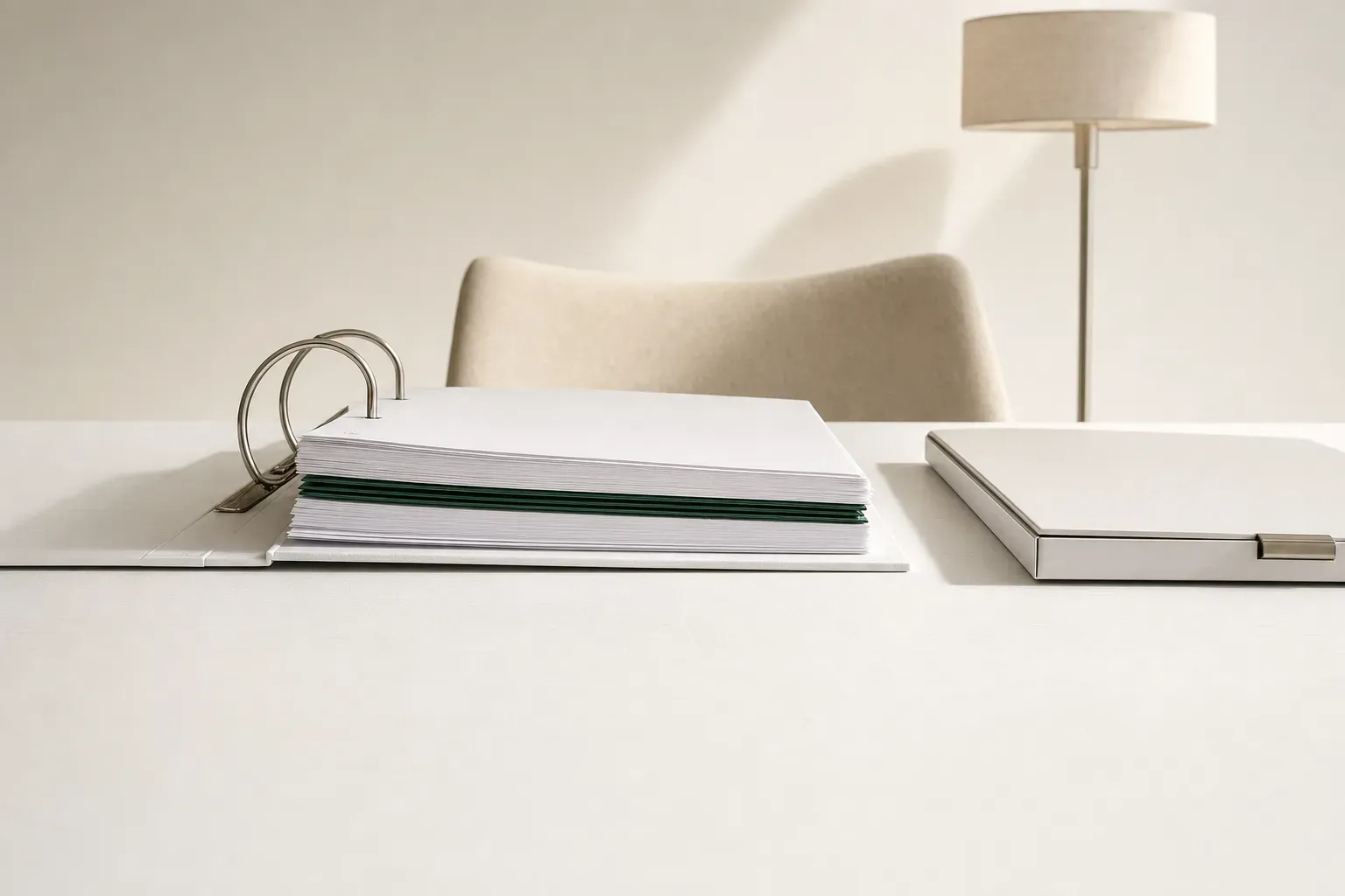 Seen low across a painted white timber dining table: an open binder of plain white printed sheets with three deep-green sheets buried part-way down the stack, one closed clasped folder beside it, an empty chair behind — what the licence file on top doesn't tell you.