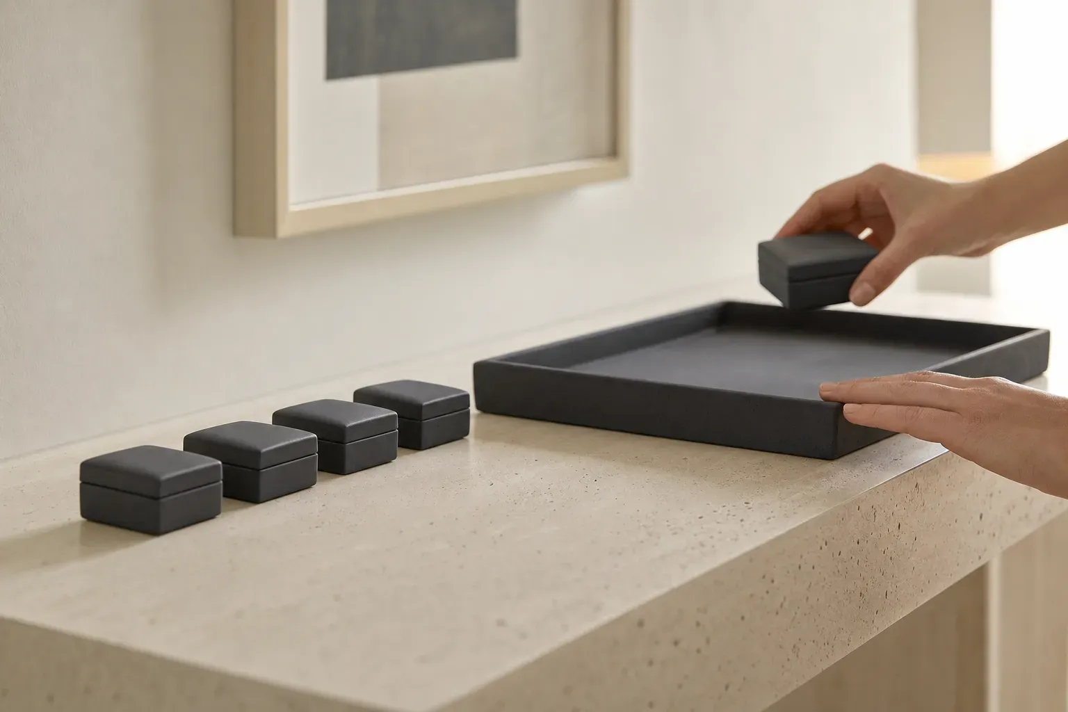 Macro close crop on a pale travertine console: five identical small charcoal ceramic boxes in a row beside one much larger empty charcoal tray big enough to hold all five, with hands at the frame edge lifting the nearest box toward it.