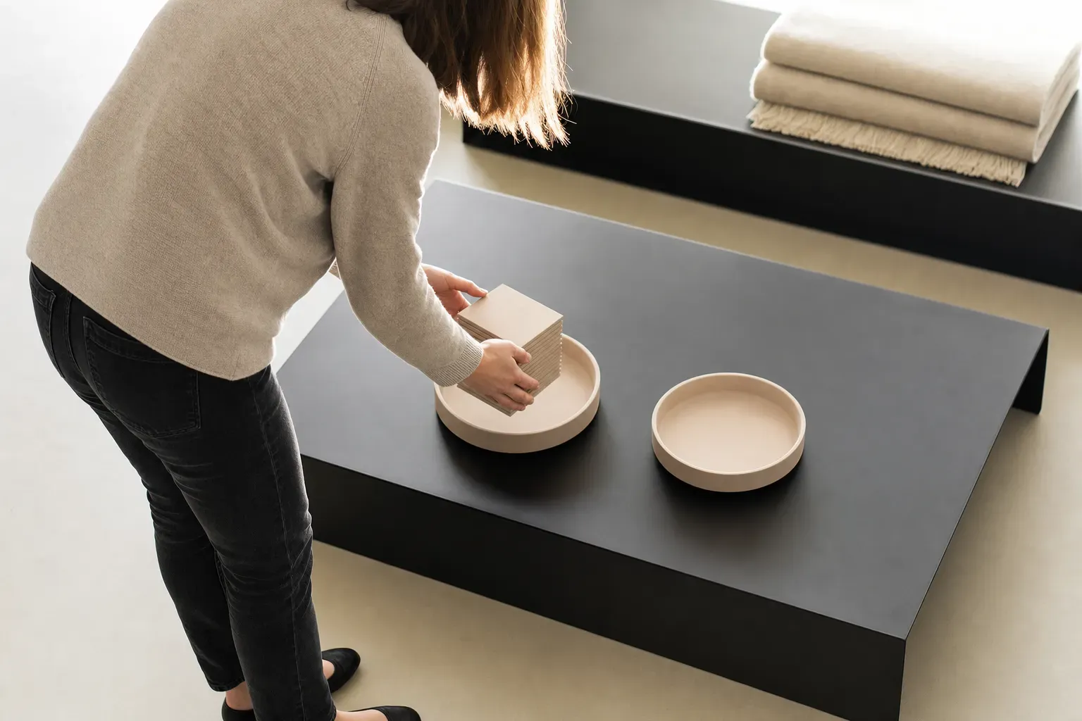 Seen from standing height at an angle onto a low matte-black steel table: two identical shallow pale-clay dishes, one holding a neat stack of small pale-clay tiles, and a standing person lifting the whole ordered set across into the empty one — moved intact, nothing left behind.