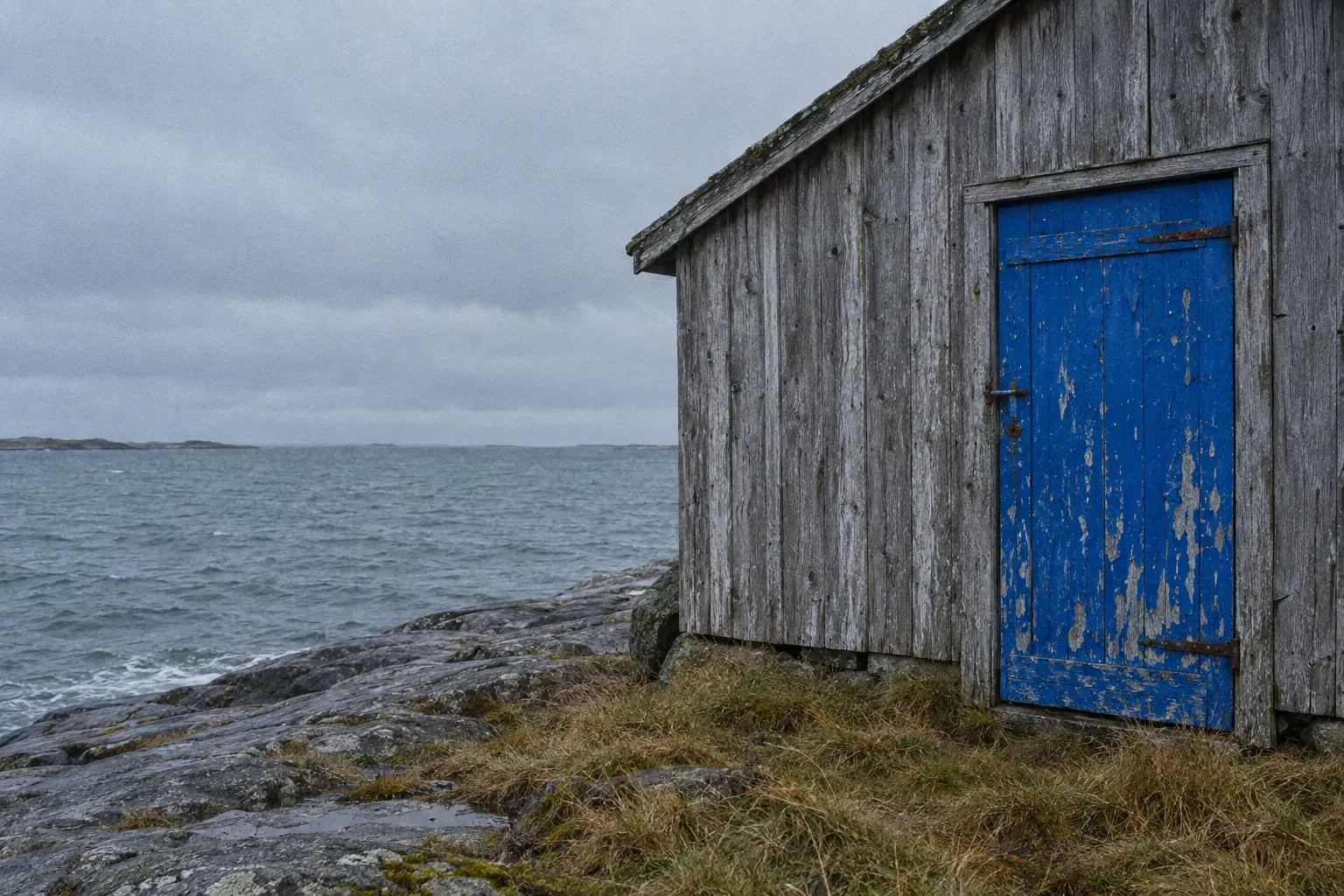 A salt-blistered cobalt blue painted door in the weathered grey plank wall of a boathouse at the water's edge, rough grass and wet granite under heavy cloud.