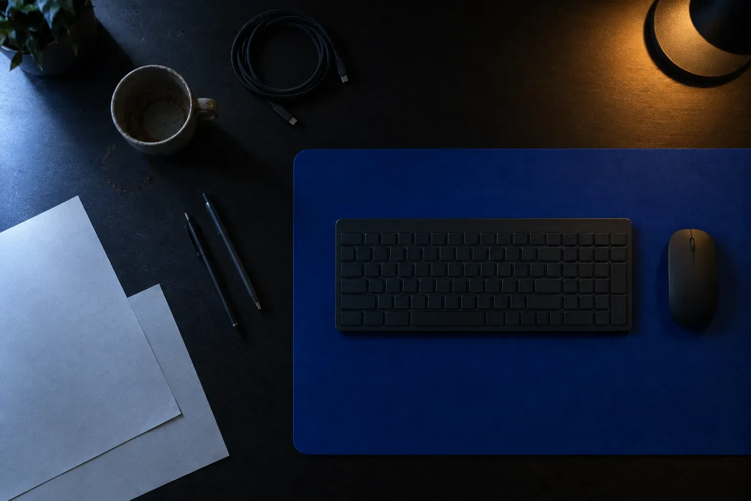 Overhead view of a dark desk at night: a large cobalt blue desk mat with a keyboard and mouse squarely on it, a mug, coiled cable and loose paper around it.