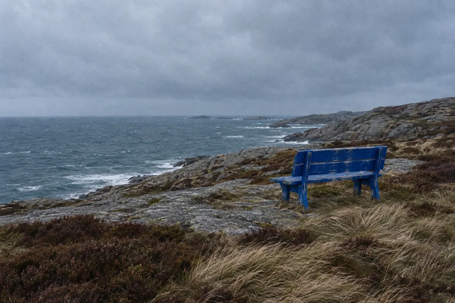 A weathered cobalt blue two-person bench standing empty on a Norwegian headland facing a grey-green sea under heavy cloud.