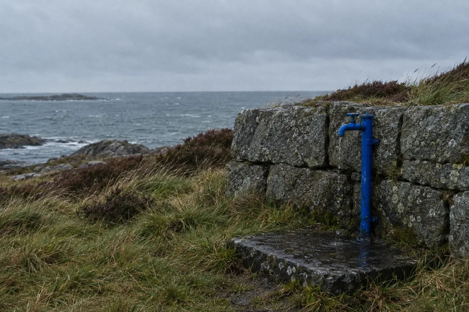 A single cobalt blue painted standpipe tap set into a grey granite wall on a Norwegian coastal campsite, a worn stone slab beneath it, cold sea beyond.