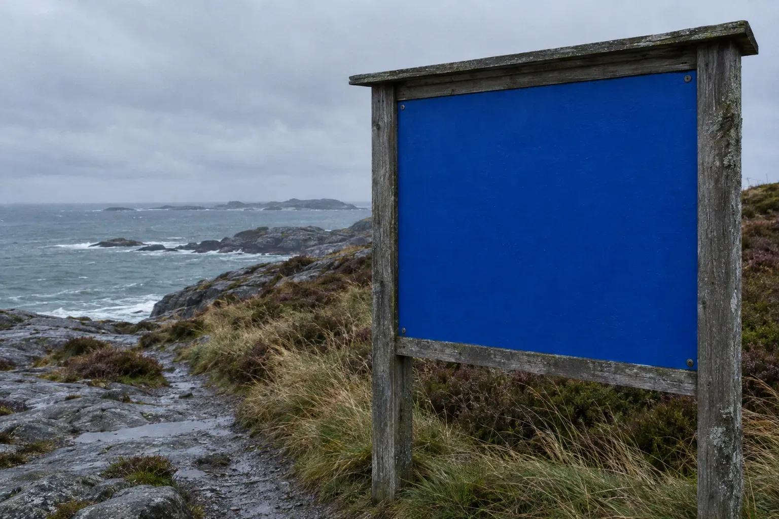 A large weathered timber-framed information board at a Norwegian coastal trailhead, its whole face painted plain cobalt blue and completely empty, cold sea beyond.