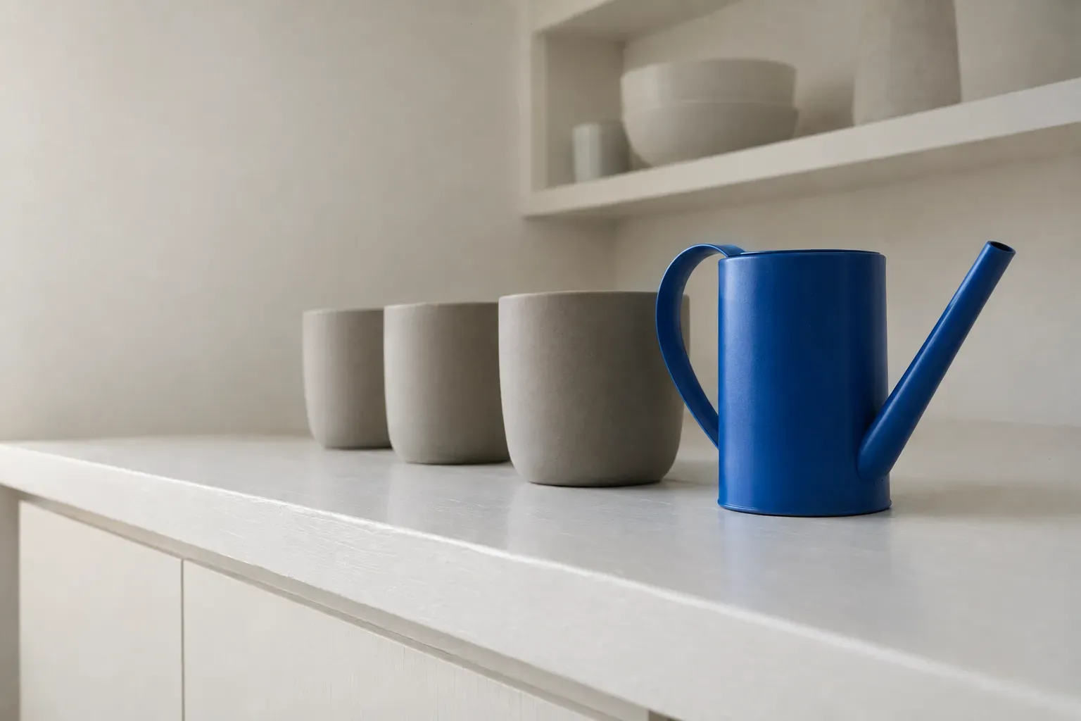 Three identical empty warm-grey ceramic pots in a row on a white-painted counter in a bright modern utility room, with one matte cobalt blue watering can standing beside them — three containers, and the upkeep that comes with owning them.
